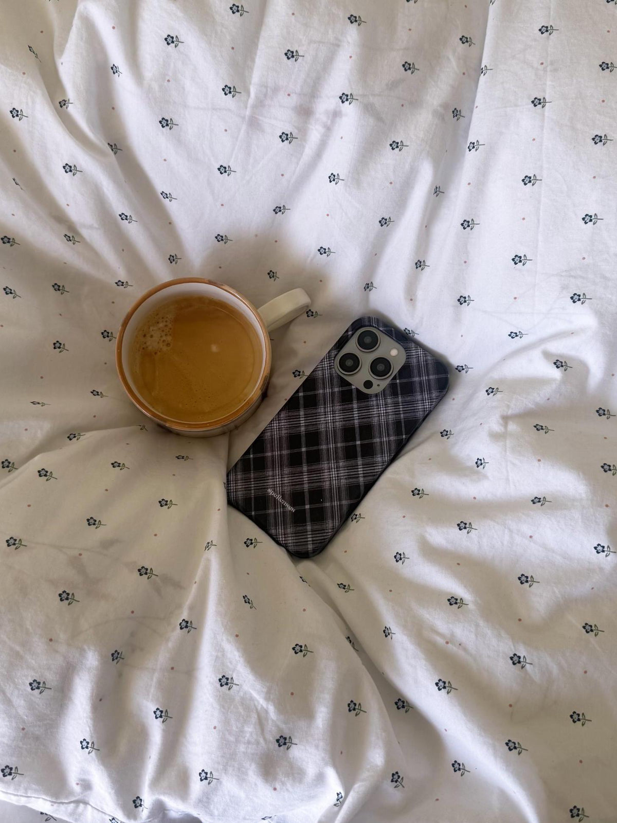 A cozy flat-lay scene on rumpled white patterned bedding, featuring a ceramic mug filled with a golden drink and a smartphone in a dark plaid case.